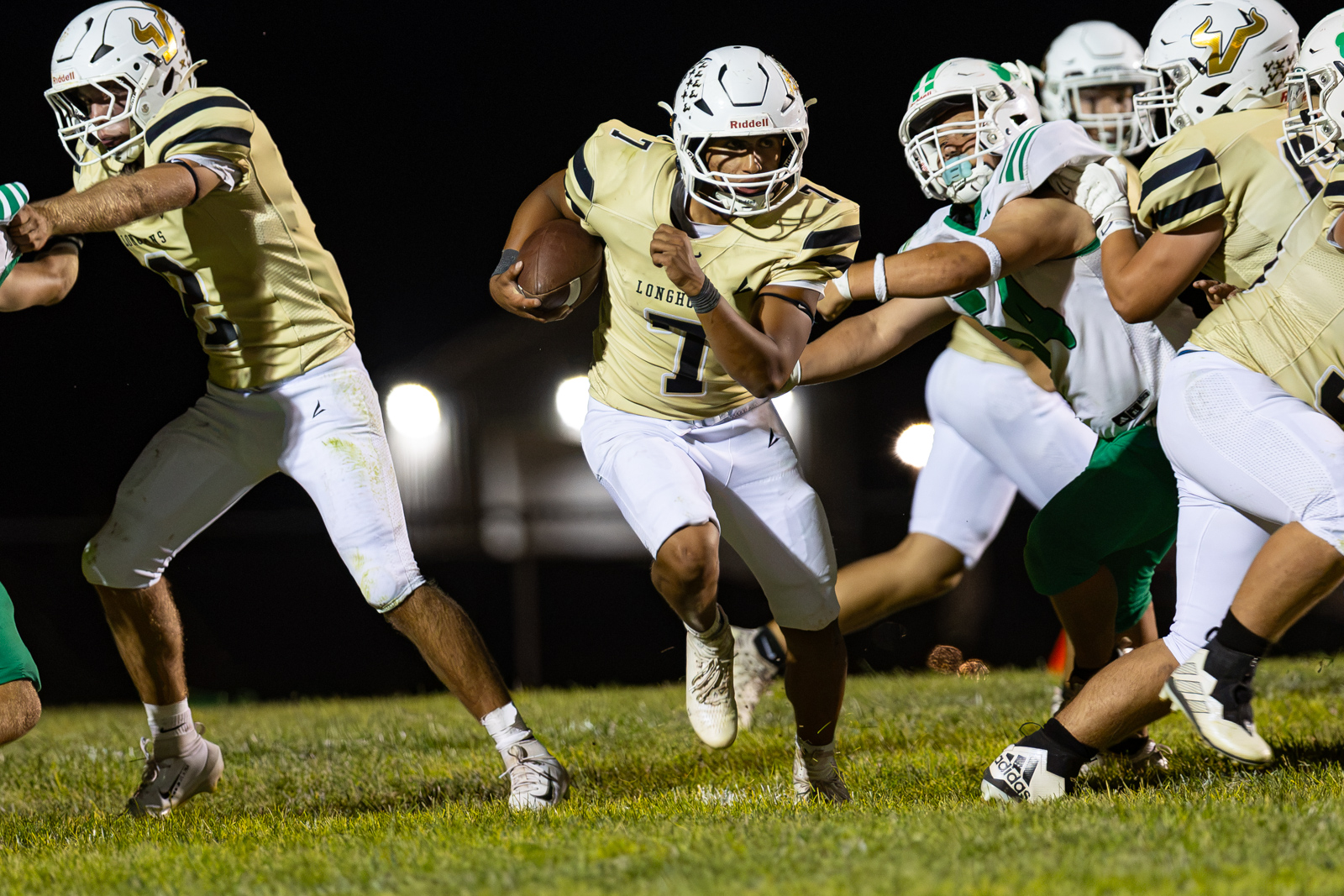 Running back in a gold jersey with the ball, sprinting forward while teammates block in a night game.