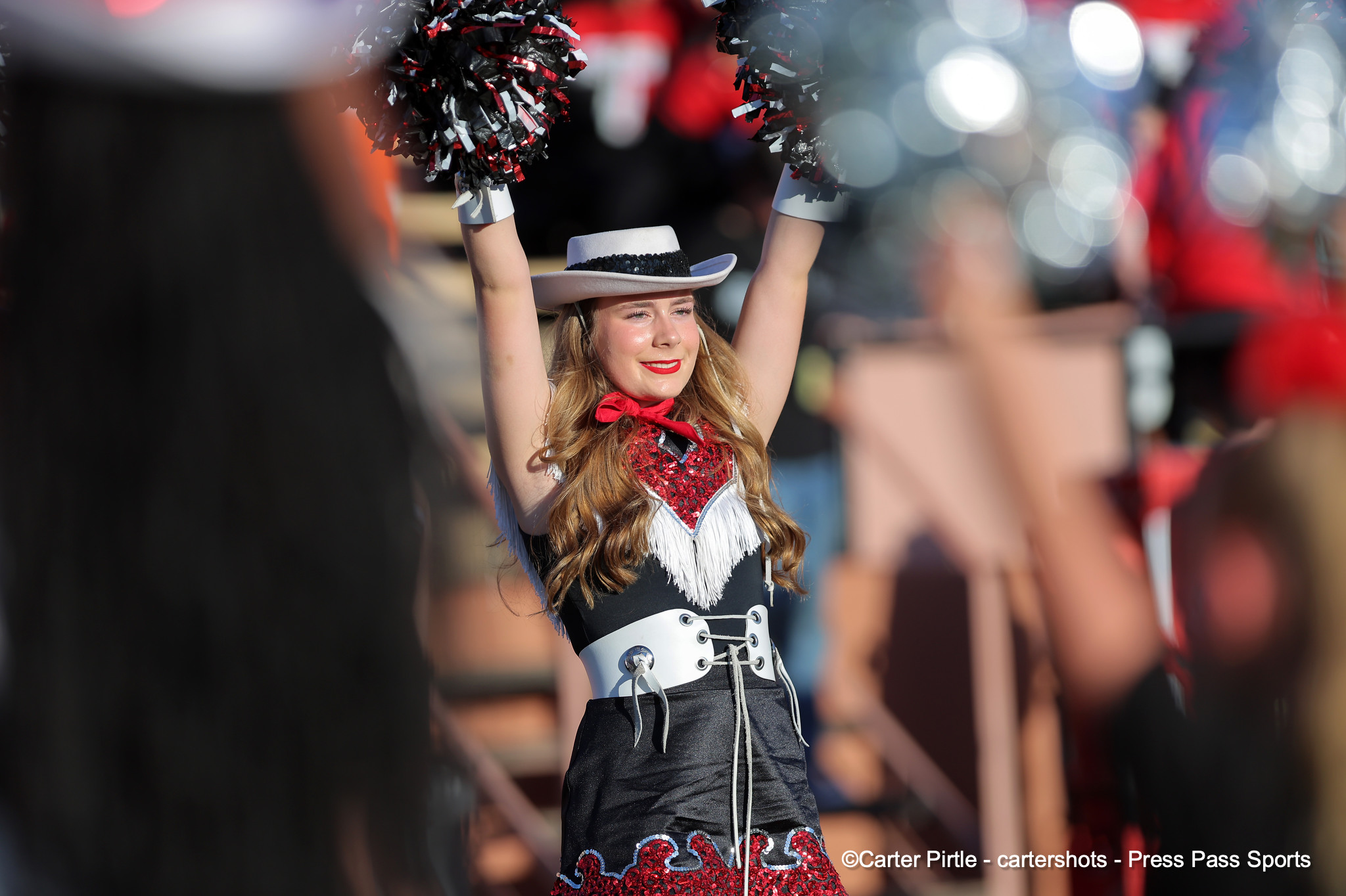Cheerleader in black-and-white uniform raises red and black pom-poms at a parade with a blurred crowd behind her.
