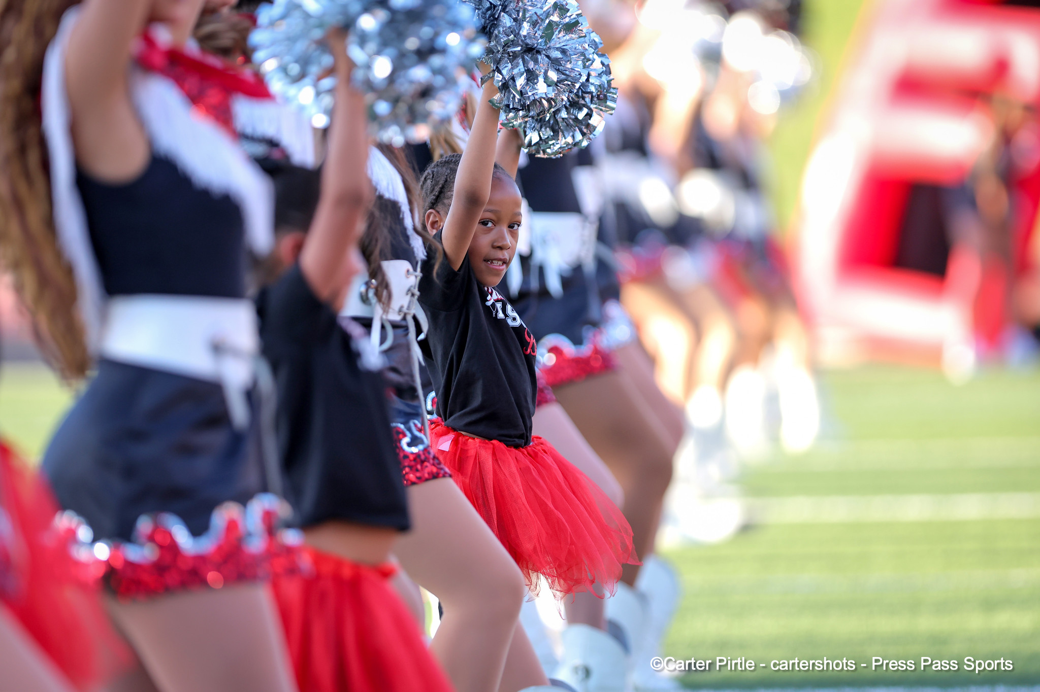 Group of cheerleaders in black tops and red skirts, raising shiny silver pom-poms on a sunny field.