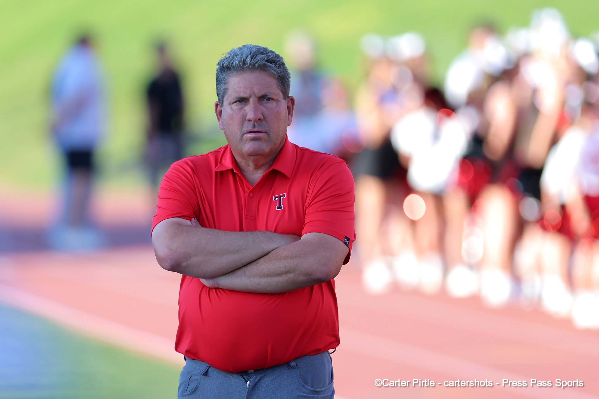 Man in a red polo with arms crossed on a running track, spectators blurred in the background.