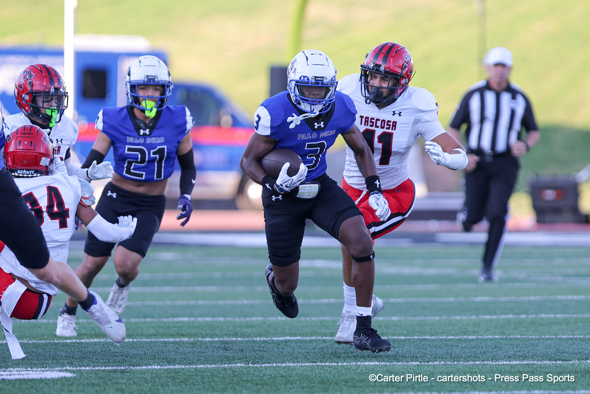 Palo Duro running back in blue carries the football, chased by a Tascosa defender in white on a football field.