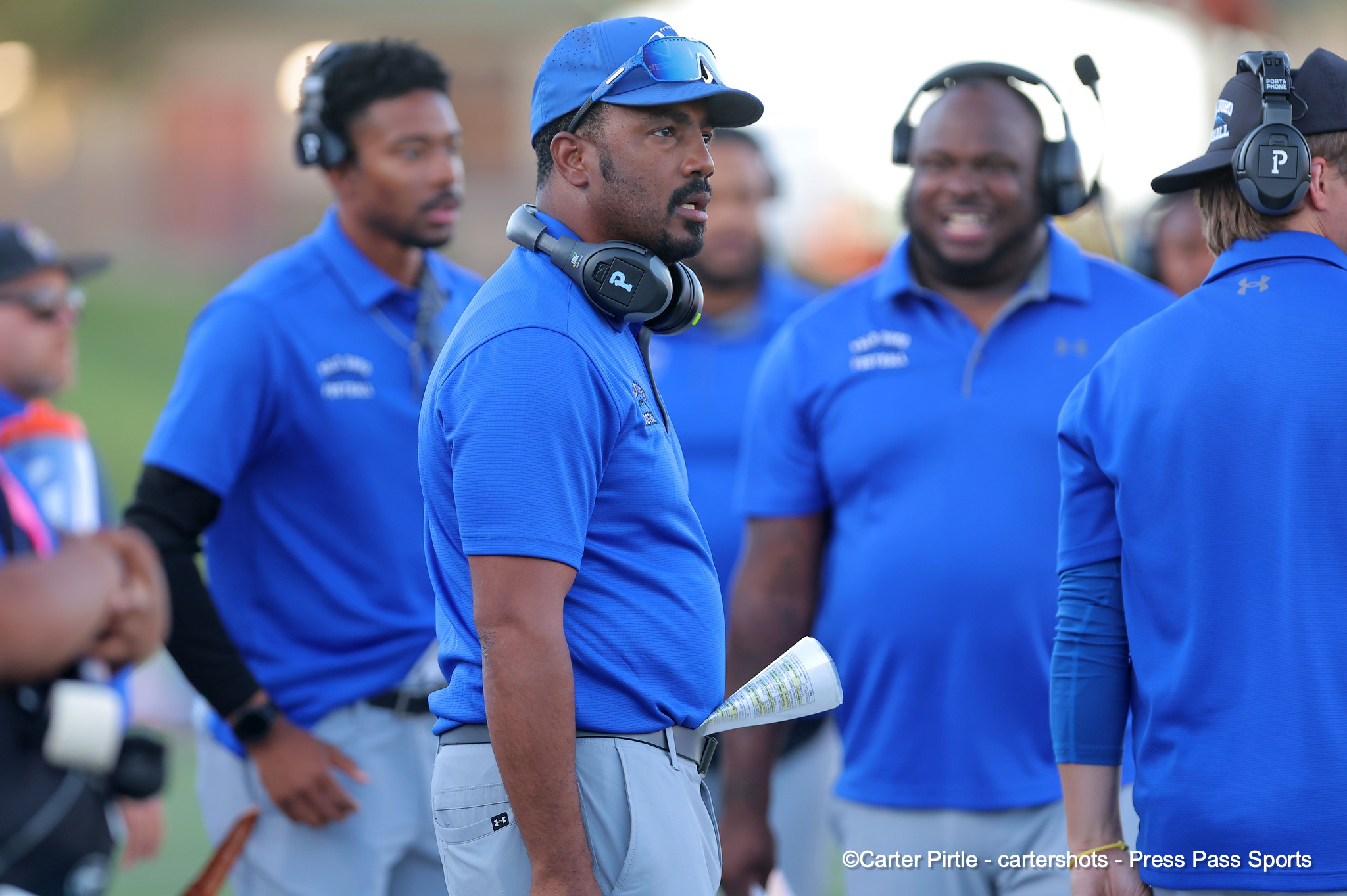 Coach wearing a blue cap and over-ear headset looks to the side while teammates in blue shirts stand around on the sideline.