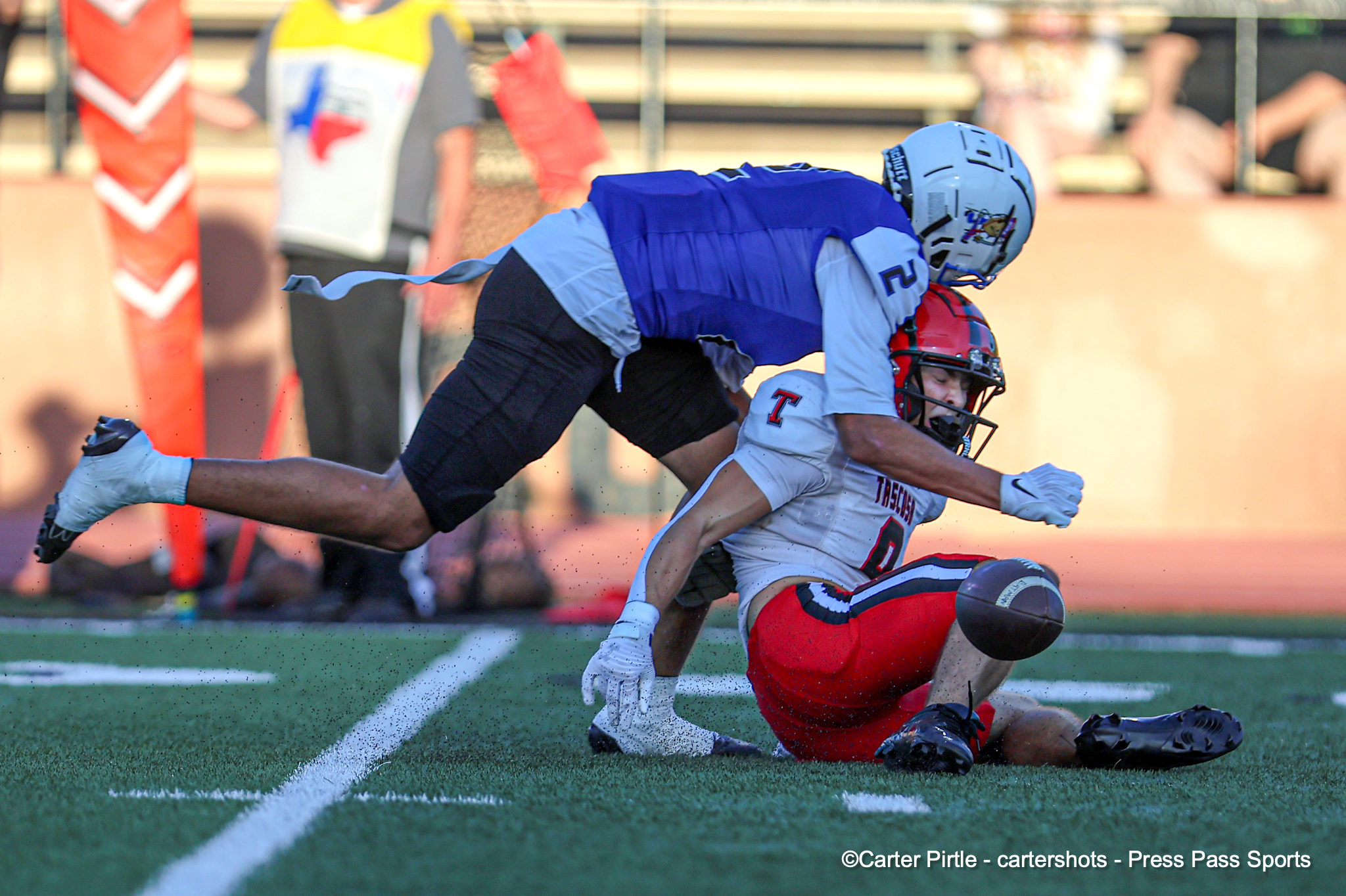 Two football players collide on a green turf field during a game, one in blue jersey tackling a white‑and‑red opponent with a red helmet.