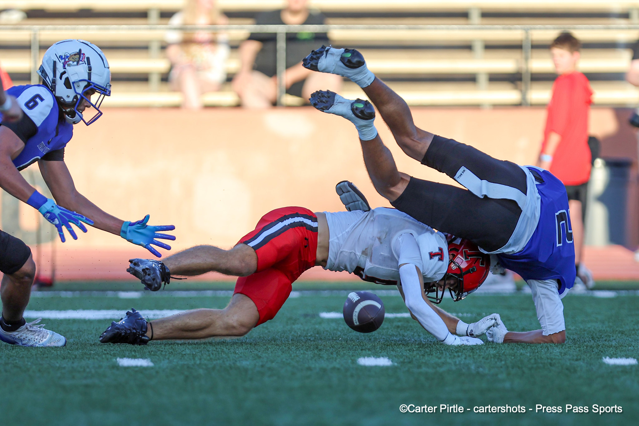 Two football players in blue uniforms collide on a green field as a ball lies on the turf nearby.