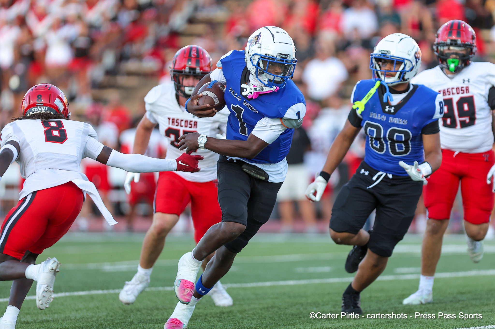 Football game action: a blue-jersey player with ball running past red- and white-uniformed defenders on a green field.