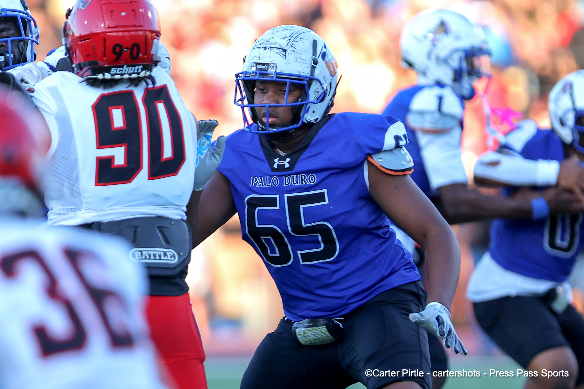 Football player in blue jersey #65 from Palo Duro collides with an opponent in a white jersey (#90) during a game on the field.