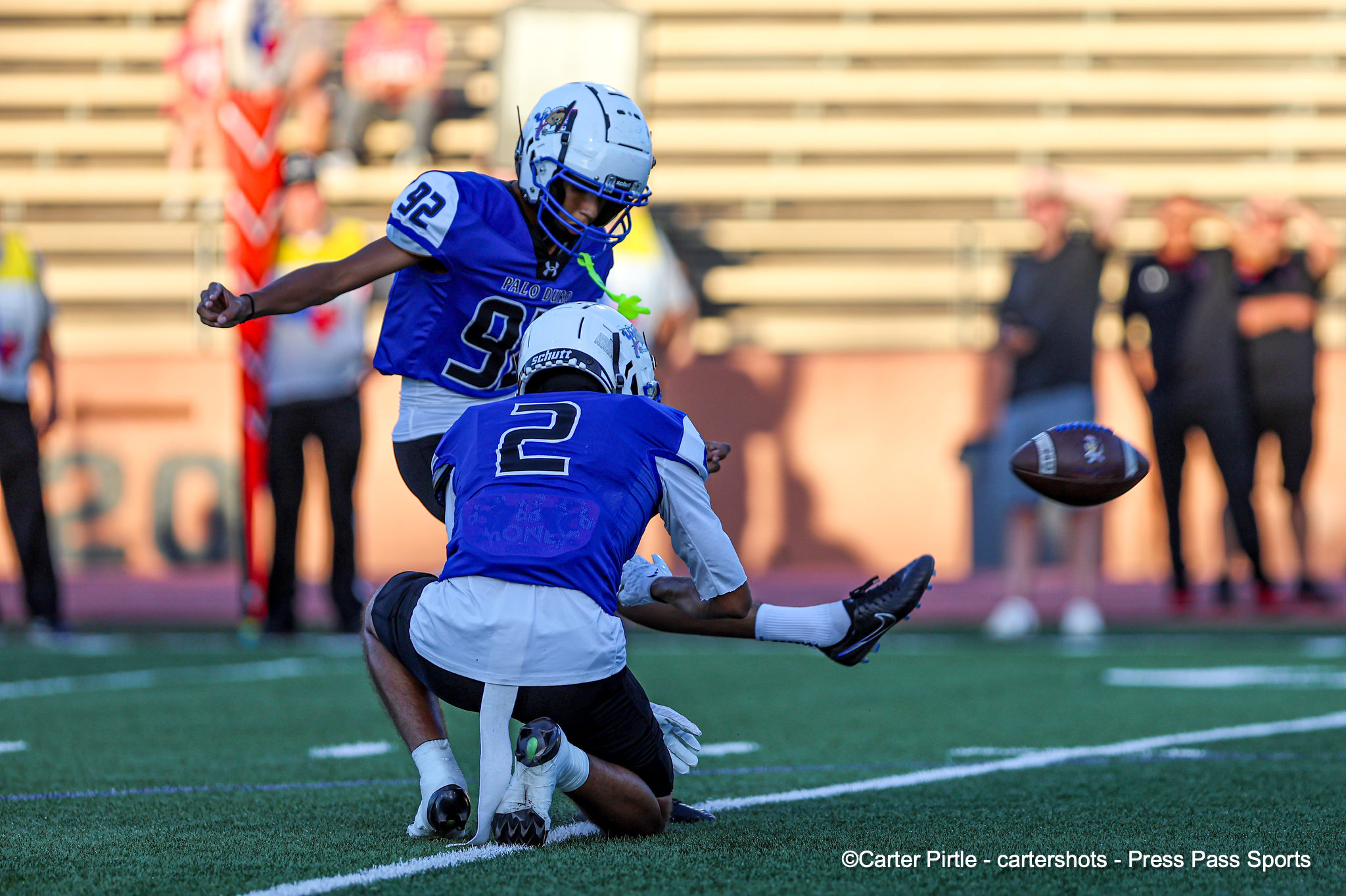 Two football players in blue uniforms collide on a grassy field, a ball rolling nearby as spectators watch in the background.