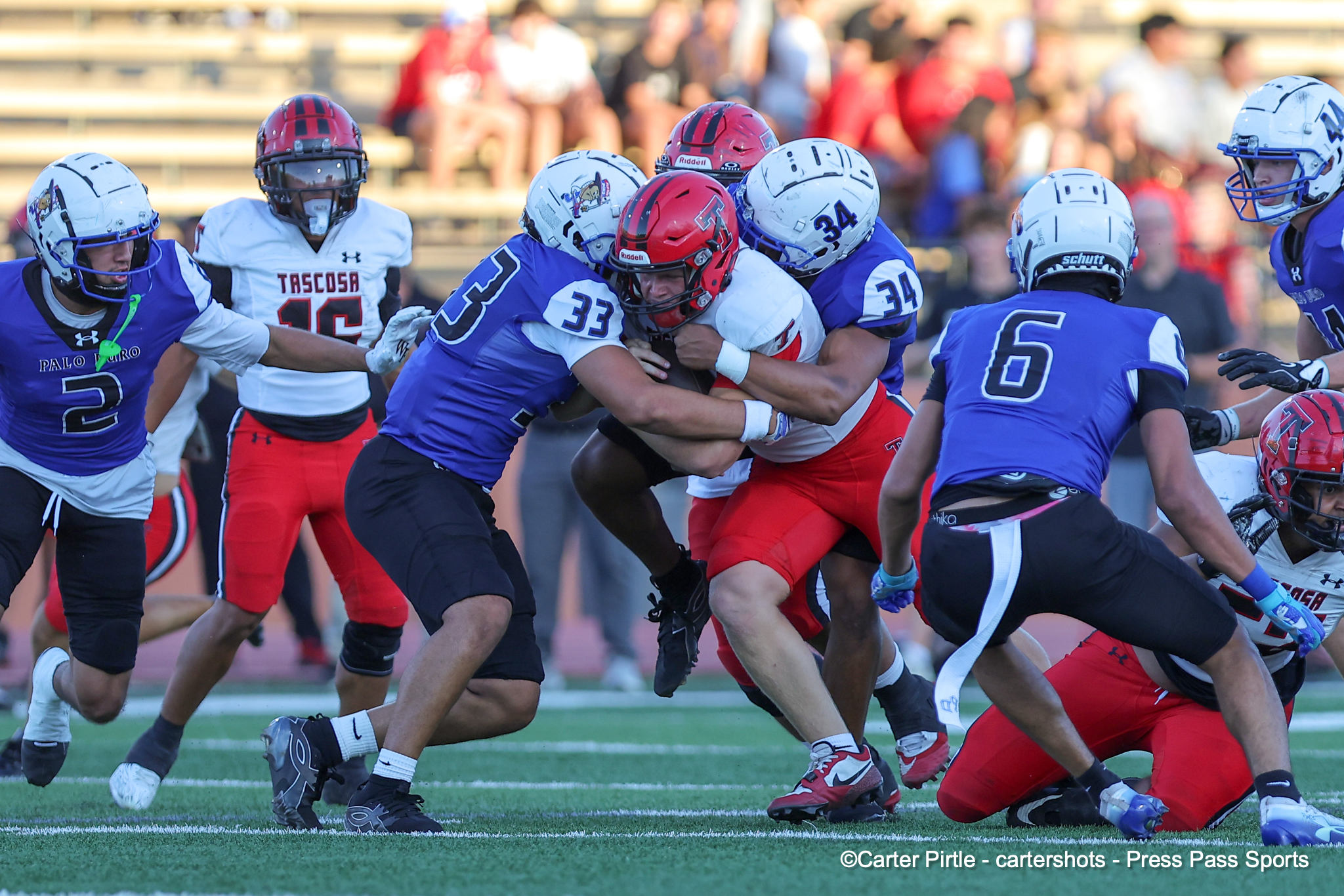 Group of football players in blue and white tackle a ball carrier in red and white on a sunny field.