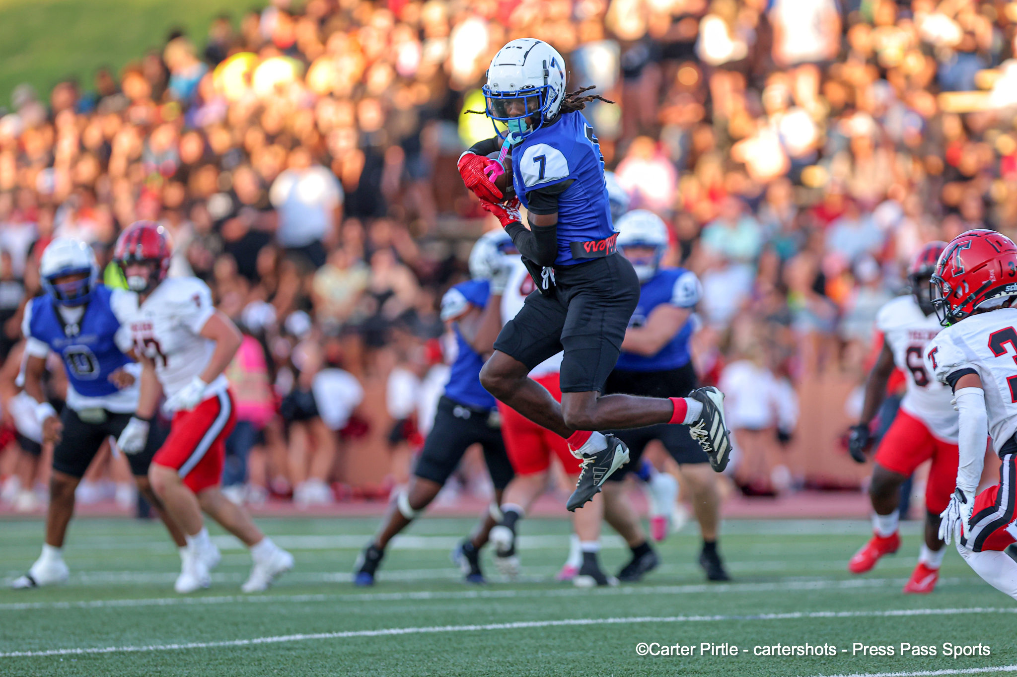 A football player in a blue jersey jumps in the air to run with the ball during a game, with a large blurred crowd in the background.