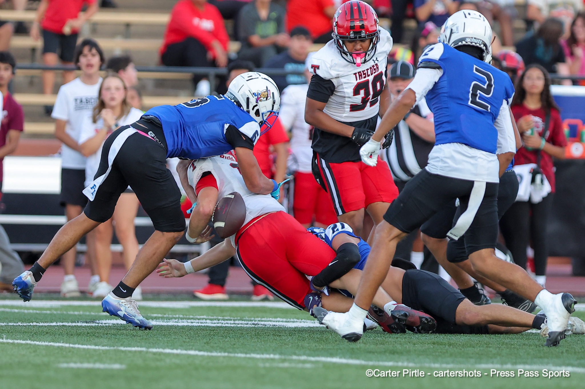 Football players in white, red, and blue uniforms collide as a ball is wrestled on a grassy field during a game; crowd watching in bleachers behind.