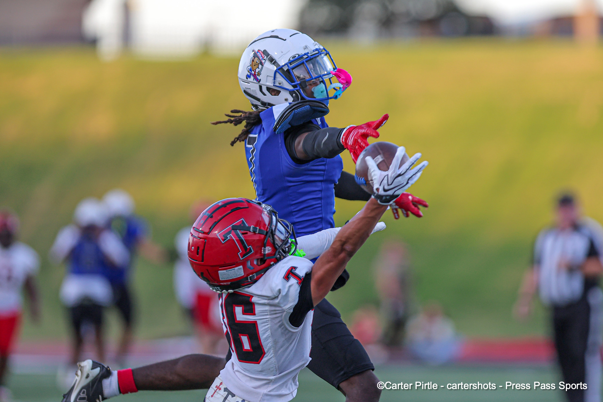 Two football players leap for the ball mid‑air; a blue‑jersey defender reaches toward a catch by a white‑jersey opponent wearing a red helmet on a grassy field.