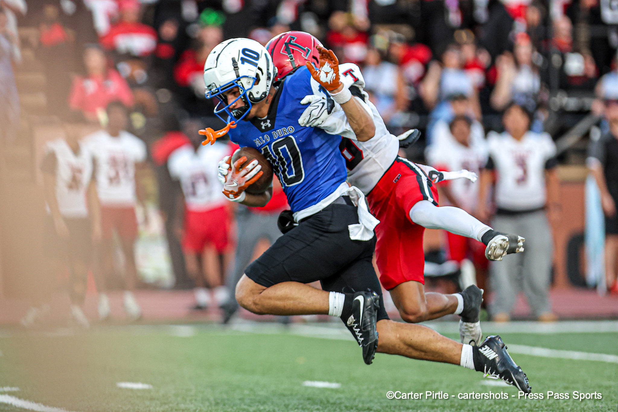 A football player in a blue jersey with the ball is tackled by a defender in white and red as a crowd watches from the stands.