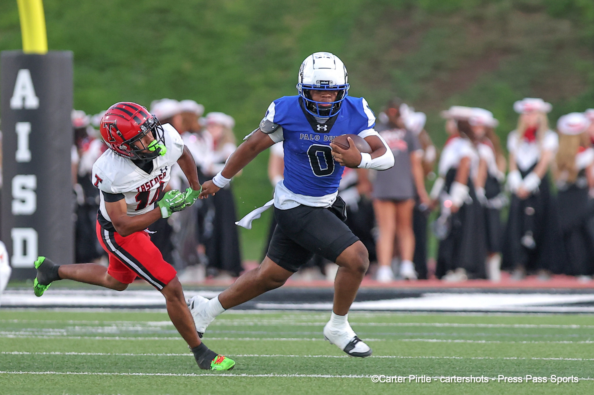 Two football players chase a ball-carrier on a green turf field; a blue-jersey runner with the ball is pursued by a white-and-red defender.