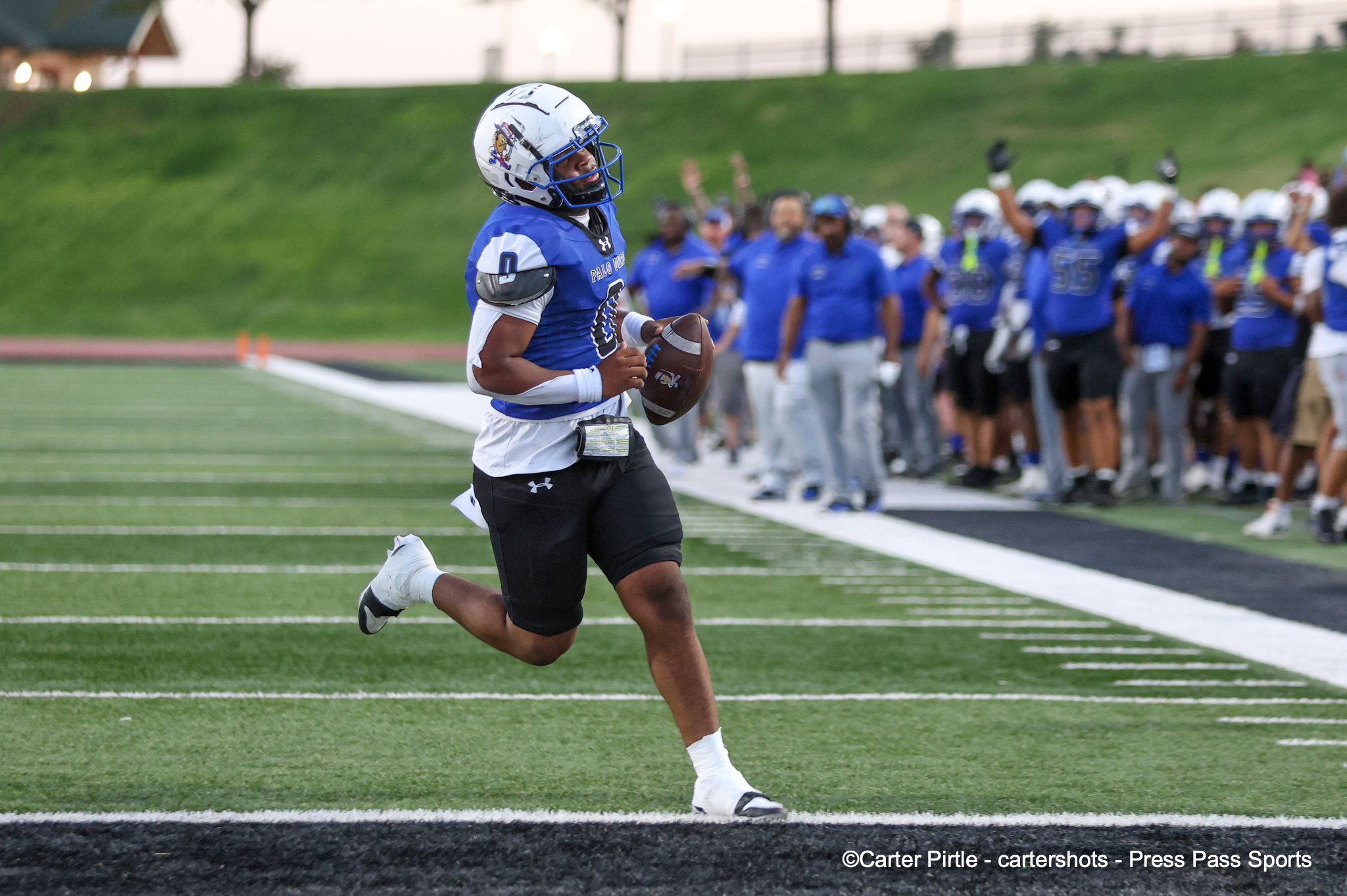 Football player in blue jersey sprinting with ball toward the end zone on a green turf field; teammates and crowd in blue uniforms cheer in the background.