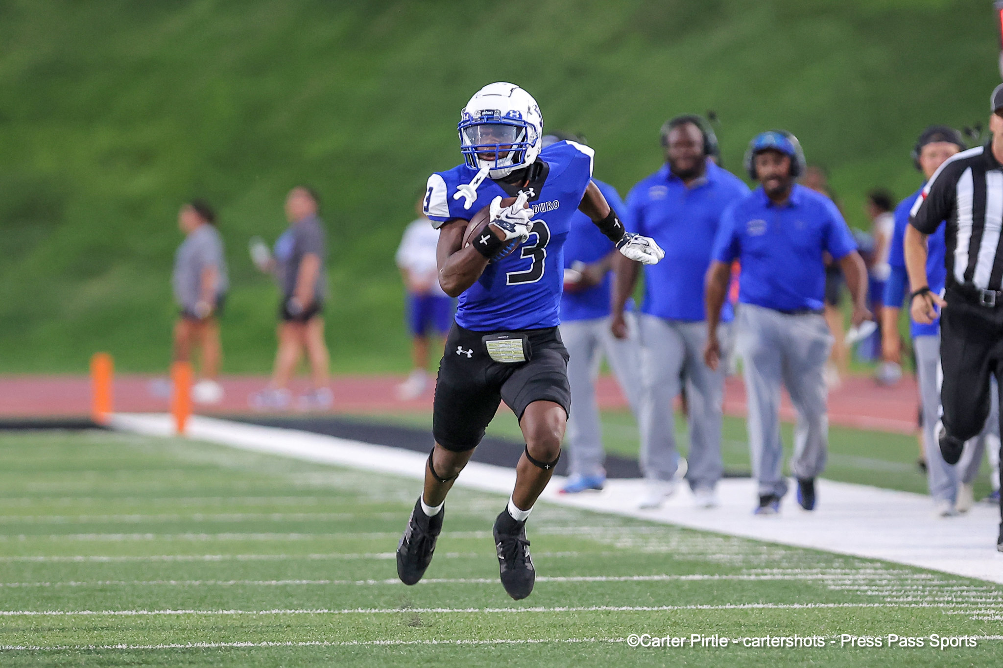 Football player in a blue uniform (number 3) runs with the ball during a game on a green turf field, with teammates and officials in the background.