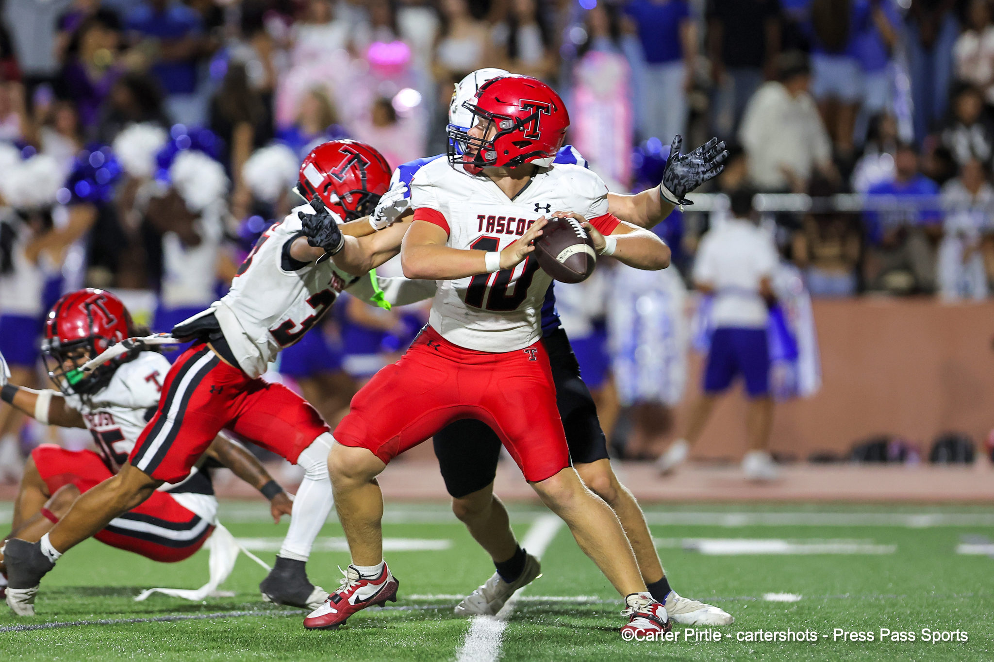 Football player in white and red uniform holds the ball and runs, pursued by teammates and opponents on a green football field with a blurred crowd in the background.
