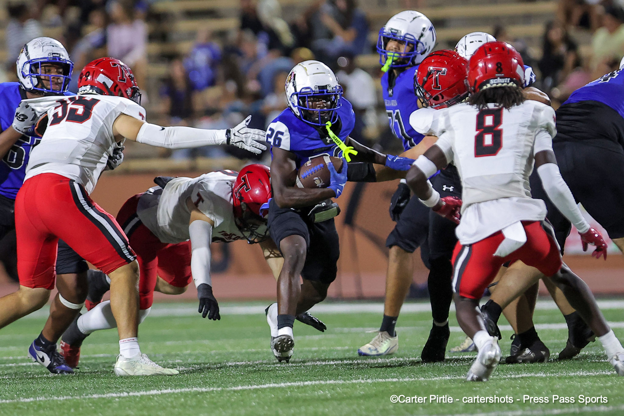 Football players in blue and white clash with white-red players as a ball carrier advances up the field.