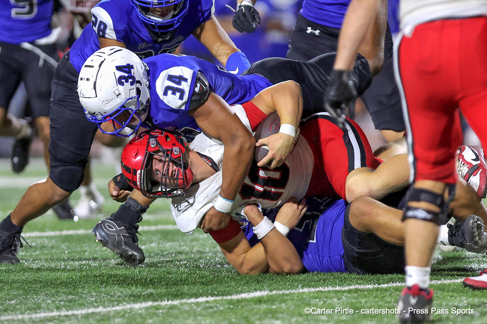 Football players in blue uniforms tackle a red-helmeted opponent on a turf field while scrambling for the ball.