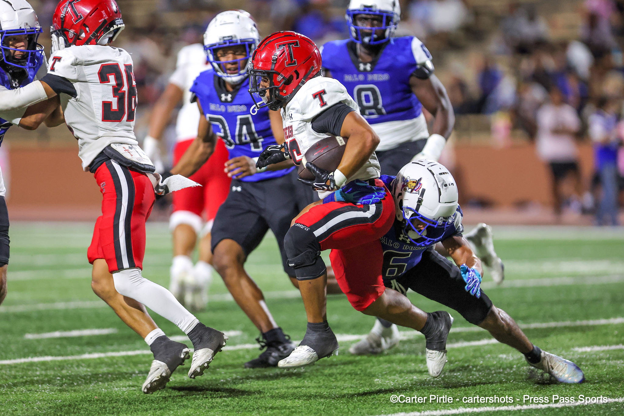 Football action: ball carrier in white and red with a red helmet runs while blue-uniform defenders close in on a green turf field.