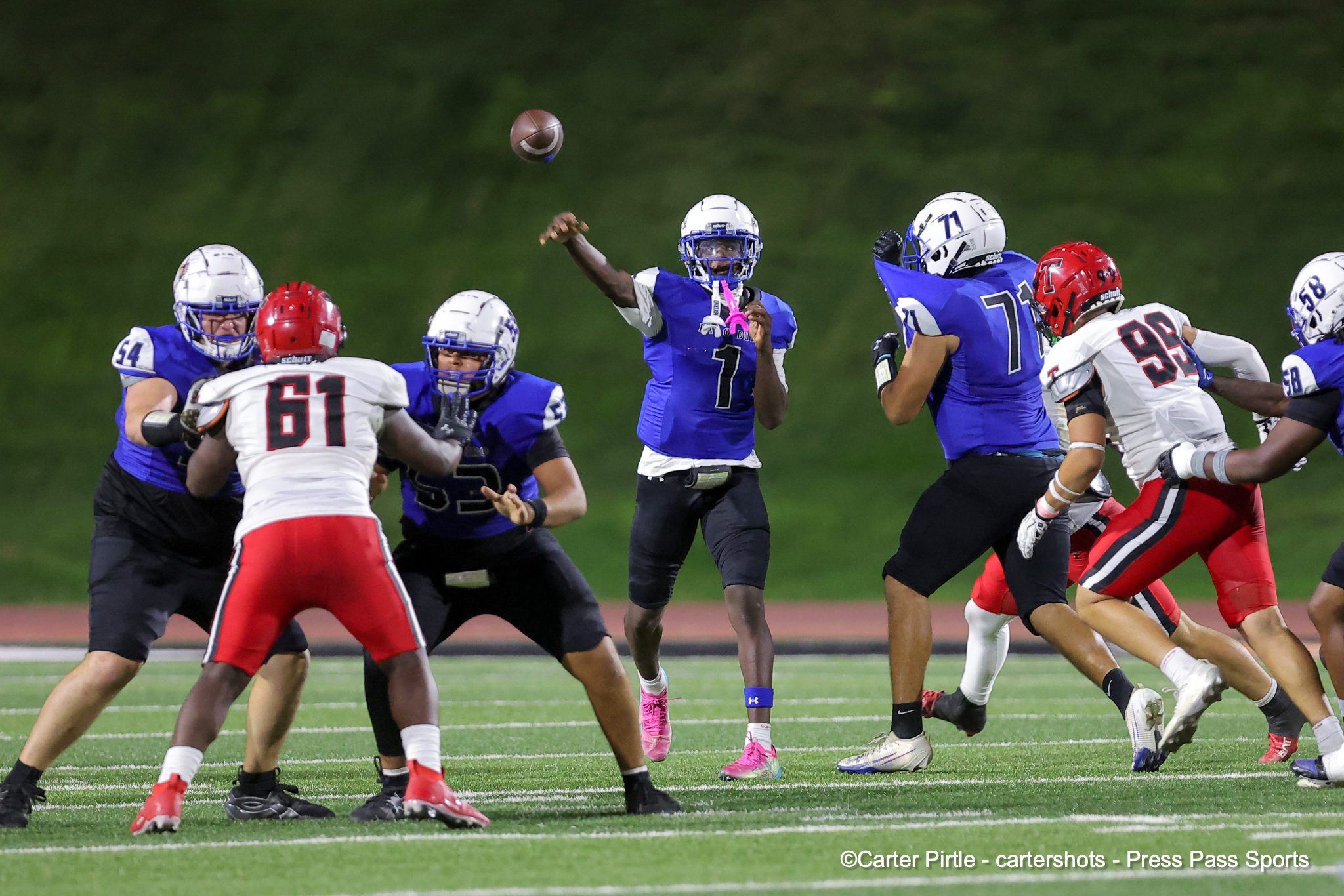 Football players in blue and white uniforms contest a pass as the ball flies midair on a bright field at night.
