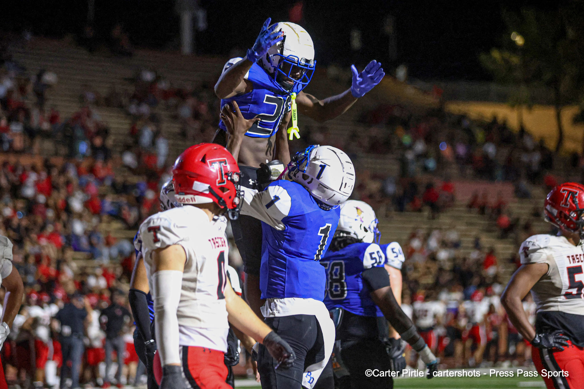 Football players in a nighttime game; a blue-team player leaps high to catch a pass while defenders reach up nearby, in a crowded stadium background.