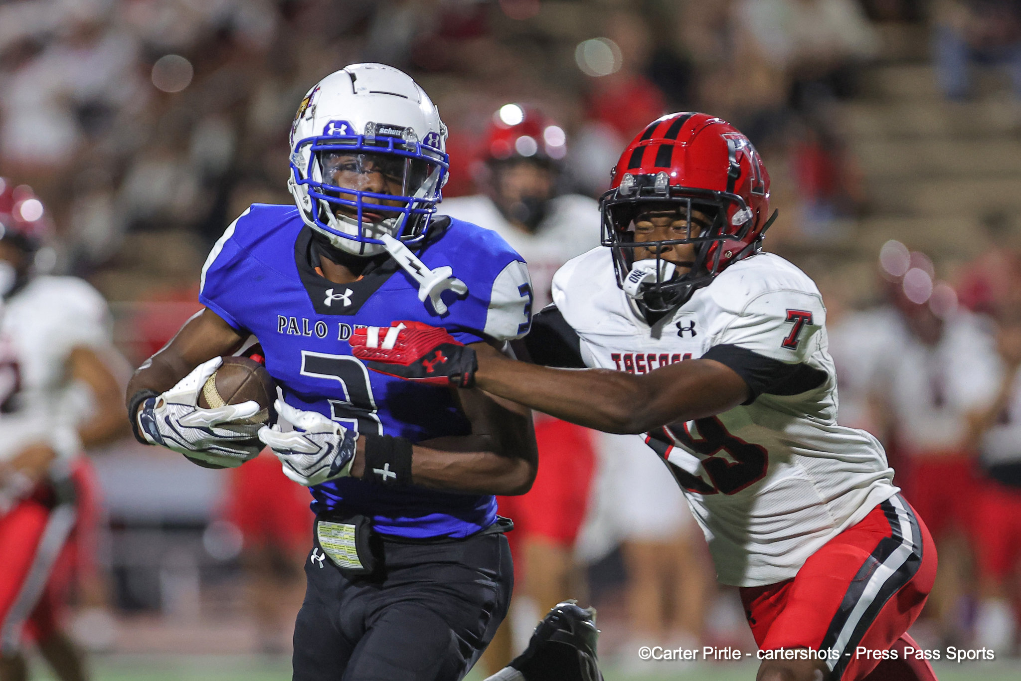Two football players in a nighttime game sprinting side by side: a blue jersey player with a white helmet (Palo D) challenges a white-jersey opponent with a red helmet for the ball.