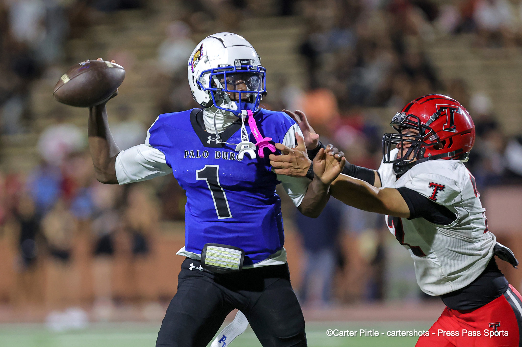 Palo Duro quarterback in blue jersey (#1) throwing a pass while a defender in red closes in.