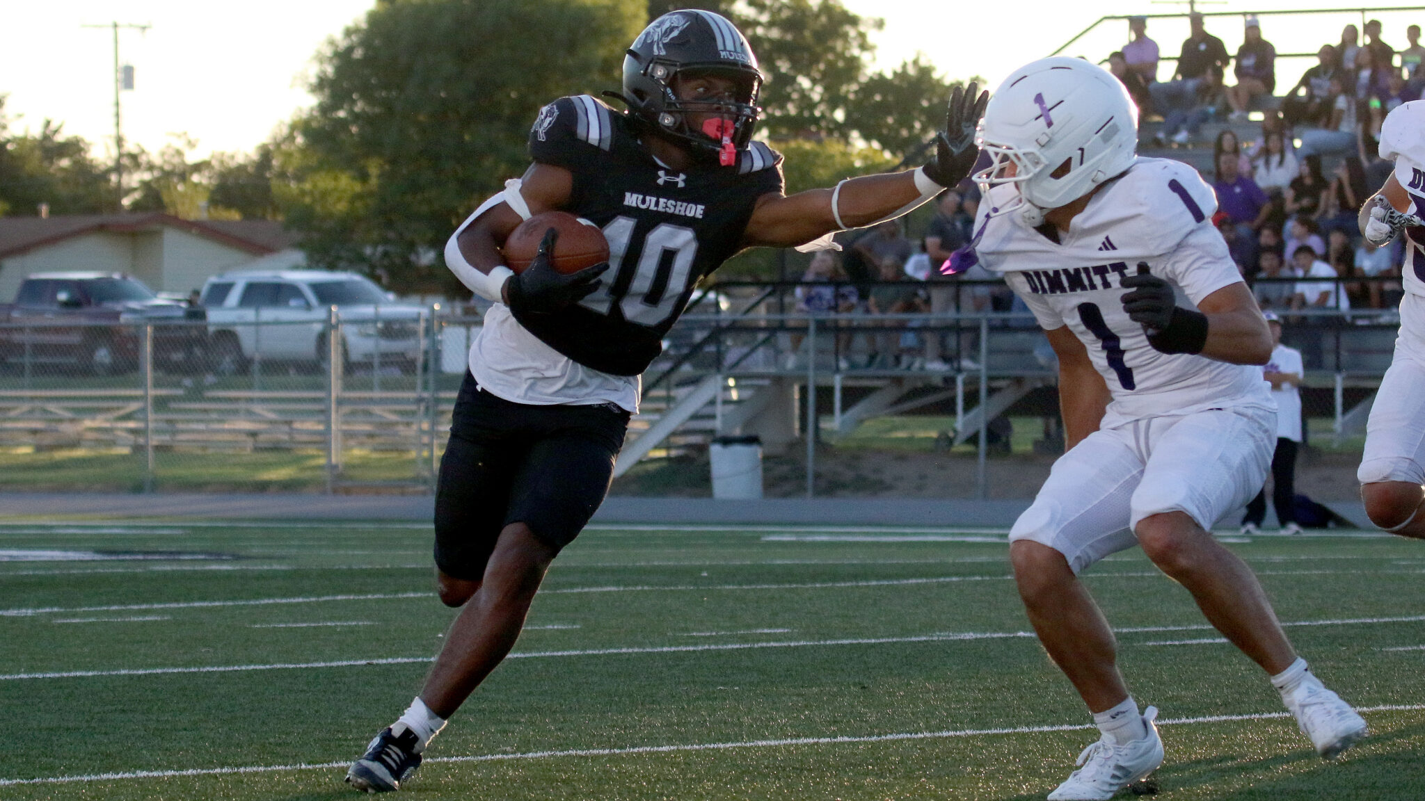 Dimmitt vs Muleshoe - Josue Segala | Press Pass Sports Football player in a black uniform carries the ball while a white-uniform defender closes in on a turf field with stands in the background.