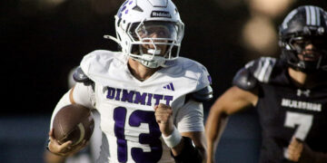 Football player in white Dimmit jersey (#23) runs with the ball, helmet on, during a game with a black-uniform opponent in the background