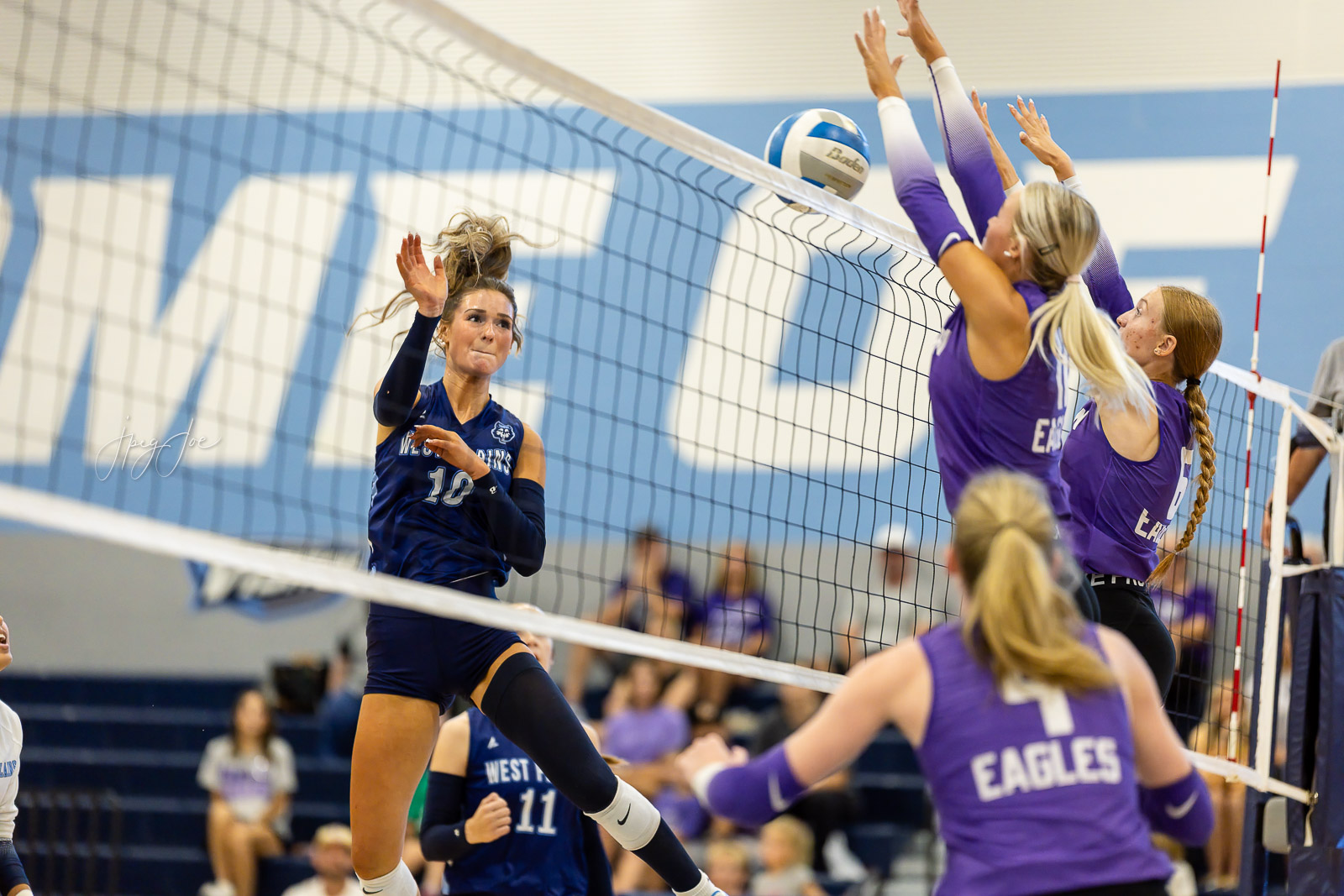 canyon-vs-west-plains-jpegJoe-volleyball-pps-8 | Press Pass Sports Blue-jersey volleyball player (#10) spikes the ball at the net as the purple team blocks defensively nearby in a gym match.