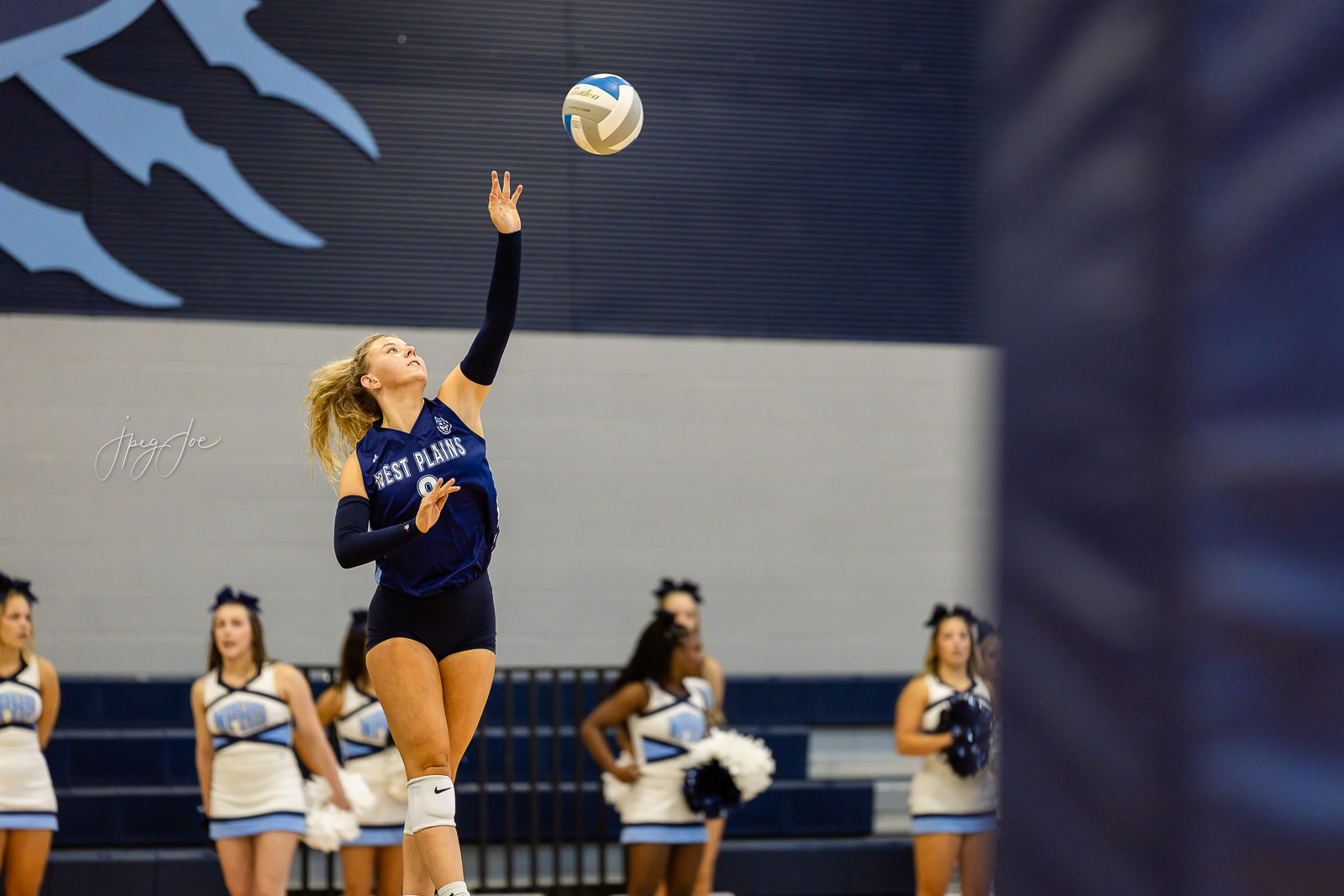 canyon-vs-west-plains-jpegJoe-volleyball-pps-10 | Press Pass Sports Female volleyball player in a blue West Plains uniform leaps to hit the ball in a gym as cheerleaders watch in the background.