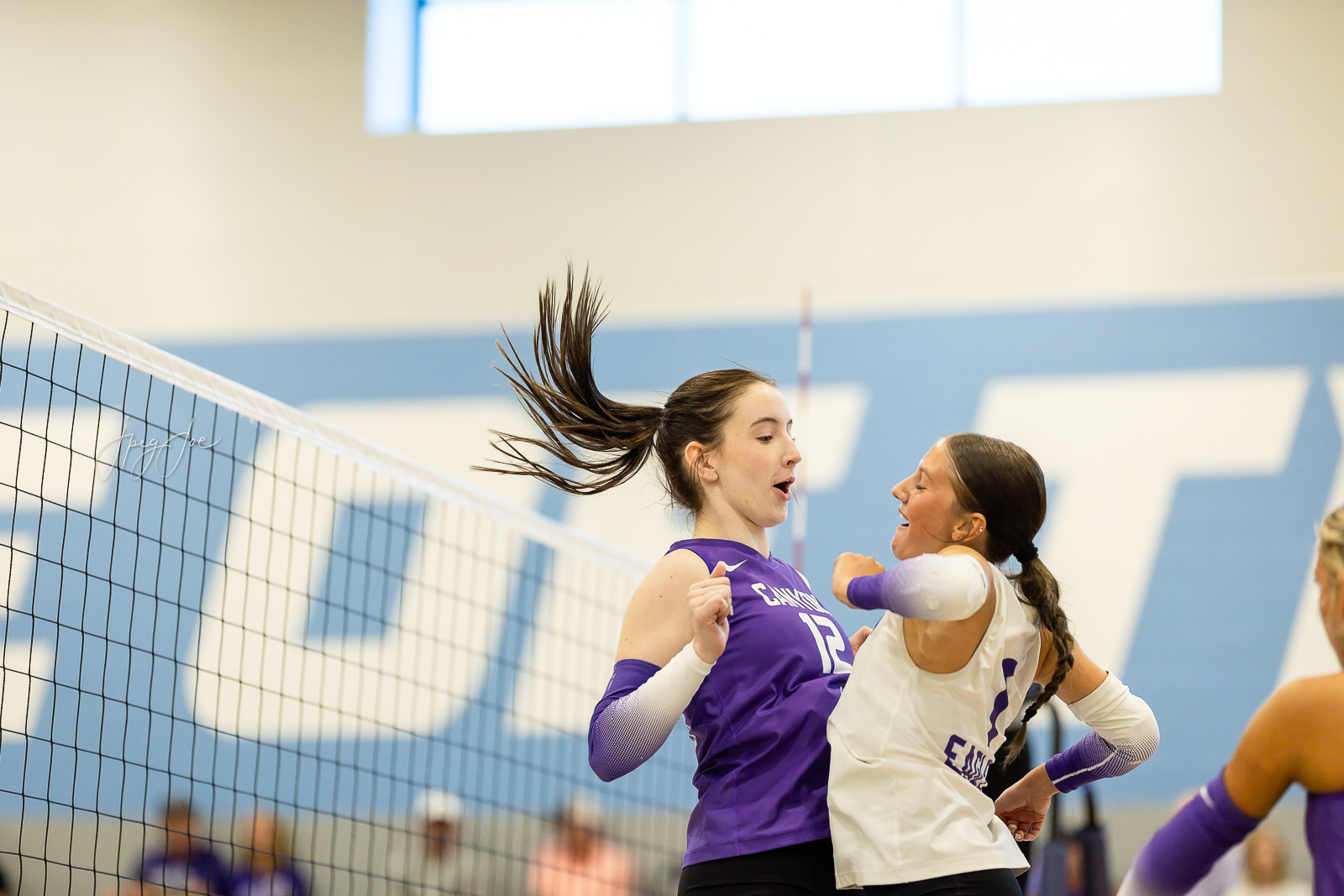 canyon-vs-west-plains-jpegJoe-volleyball-pps-16 | Press Pass Sports Two female volleyball players collide near the net during a rally, one in a purple jersey and the other in white, with hair flying mid‑air gym scene