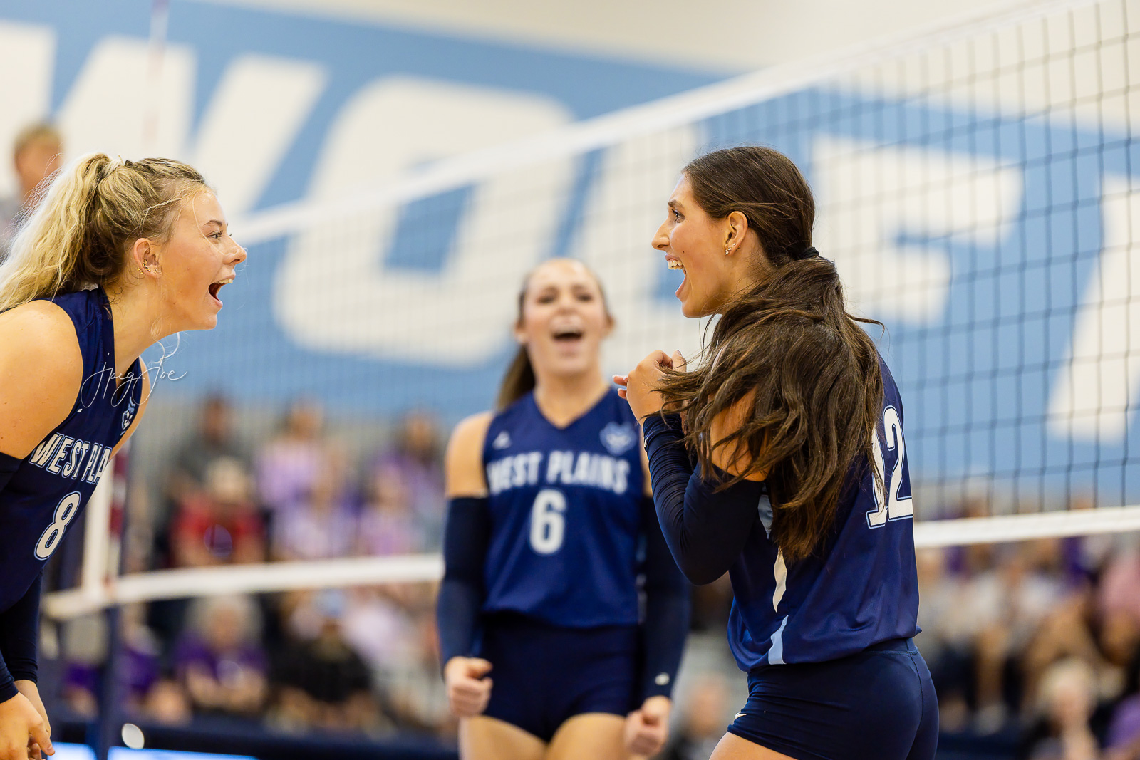 canyon-vs-west-plains-jpegJoe-volleyball-pps-17 | Press Pass Sports Three volleyball players in blue uniforms celebrate near the net during a match, faces lit with joy as they cheer together.