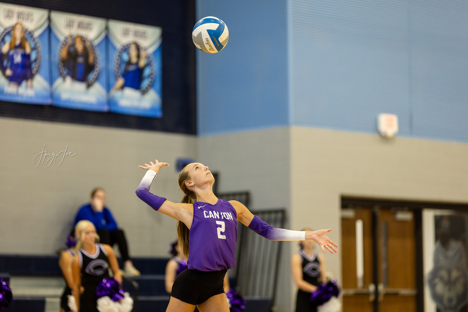canyon-vs-west-plains-jpegJoe-volleyball-pps-23 | Press Pass Sports Female volleyball player in a purple jersey (#2) leaps to serve the ball during a match, with banners and spectators in the gym background.