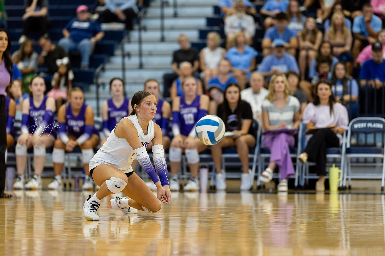 canyon-vs-west-plains-jpegJoe-volleyball-pps-26 | Press Pass Sports A female volleyball player in white dives toward the ball on a polished gym floor as spectators watch from the bleachers behind her on game day.