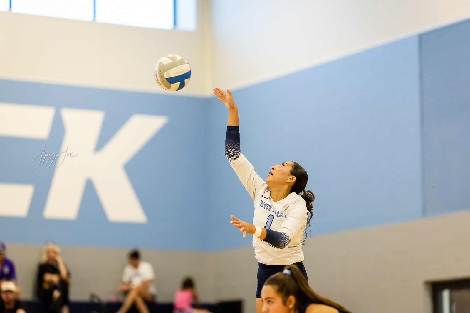 canyon-vs-west-plains-jpegJoe-volleyball-pps-27 | Press Pass Sports Female volleyball player in white/blue uniform extending arm to serve the ball in a gym with blurred spectators in the background.