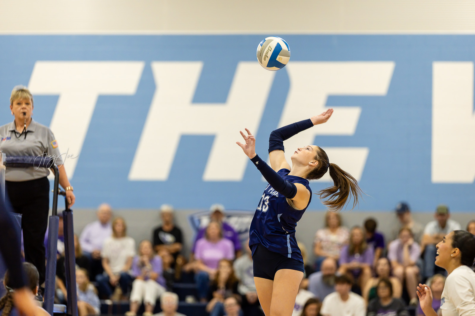canyon-vs-west-plains-jpegJoe-volleyball-pps-28 | Press Pass Sports Female volleyball player in a blue uniform serves the ball mid-air, with a referee and a cheering crowd in a gym.