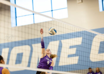 Female volleyball player in a purple jersey (#9) spikes the ball over the net in a gym.