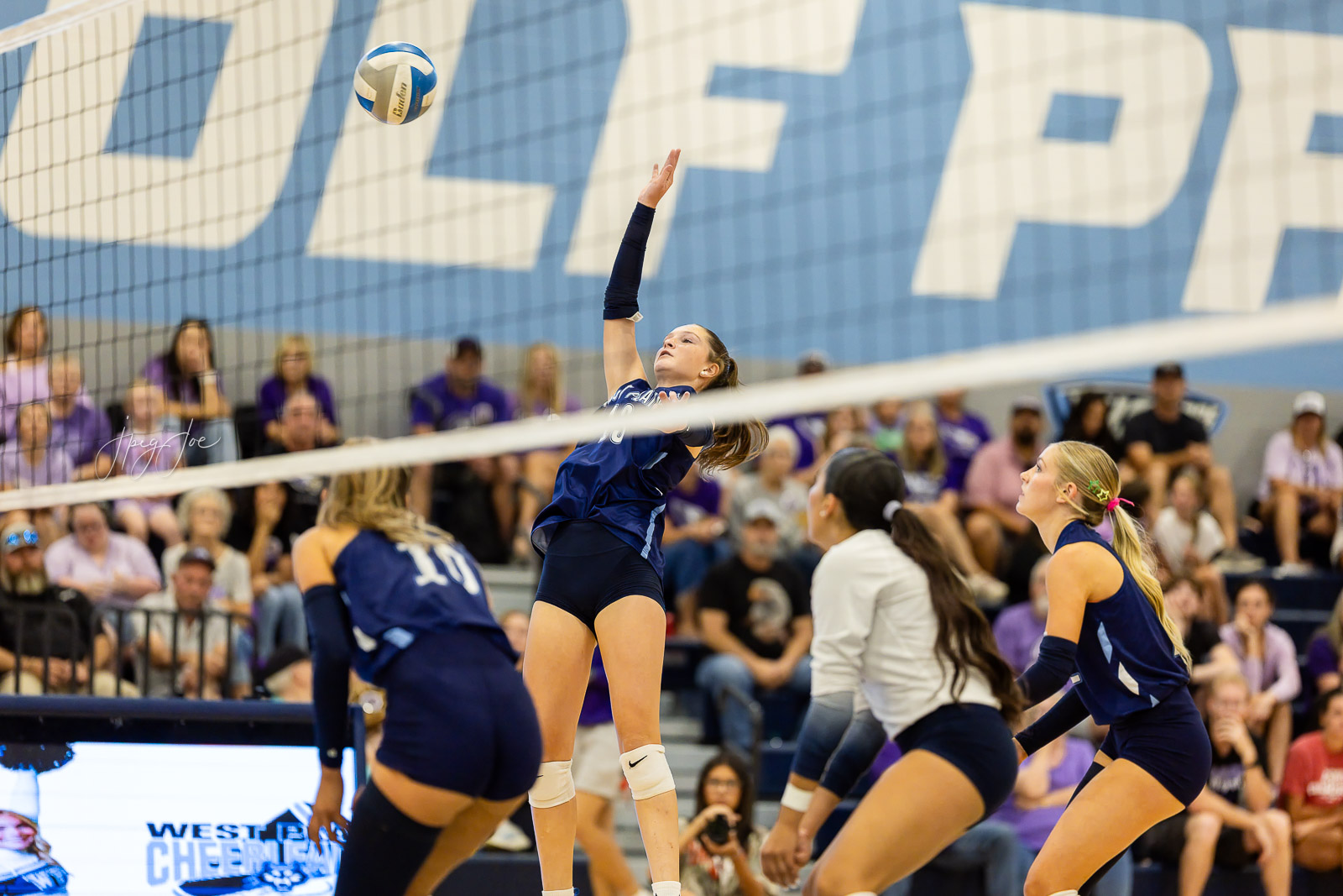 canyon-vs-west-plains-jpegJoe-volleyball-pps-32 | Press Pass Sports Female volleyball player in navy uniform jumps to spike the ball over the net as teammates and spectators watch.