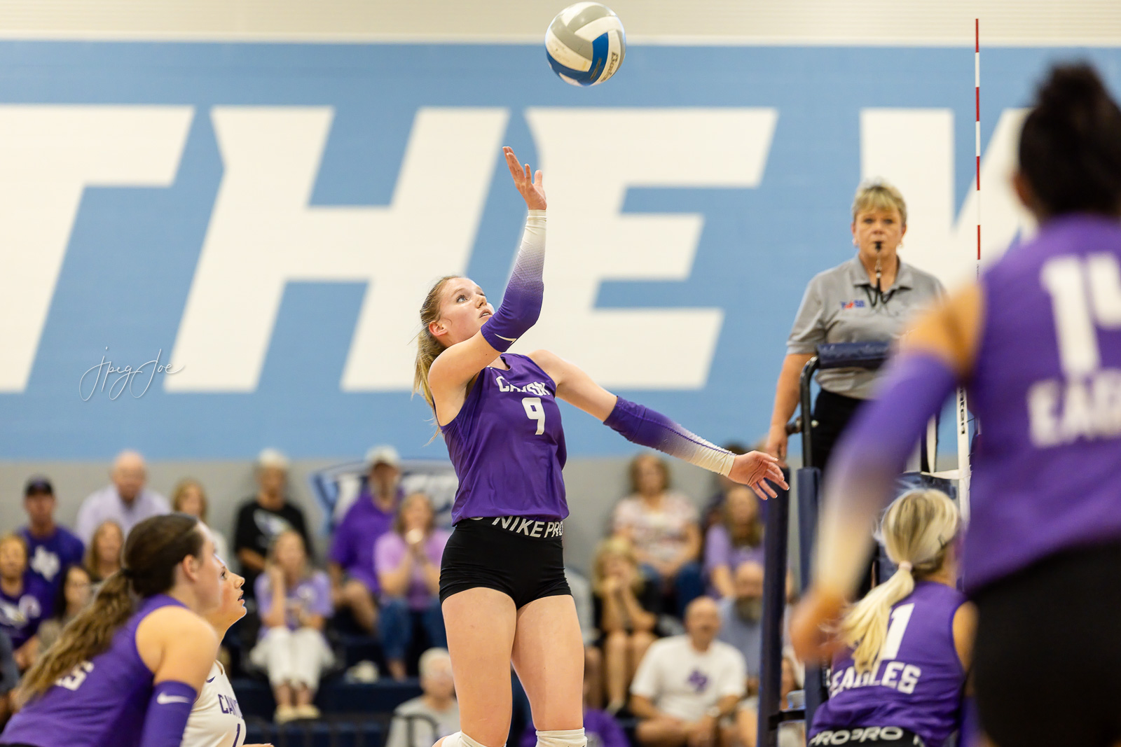 canyon-vs-west-plains-jpegJoe-volleyball-pps-33 | Press Pass Sports Volleyball player in purple jersey with number 9 serving the ball during a match in a gym with a blue wall and spectators watching.