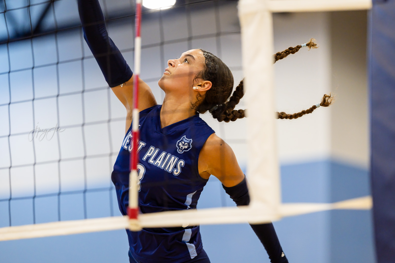 canyon-vs-west-plains-jpegJoe-volleyball-pps-34 | Press Pass Sports Female volleyball player in a blue West Plains jersey reaching up to block at the net during an indoor match, braids flying behind her.