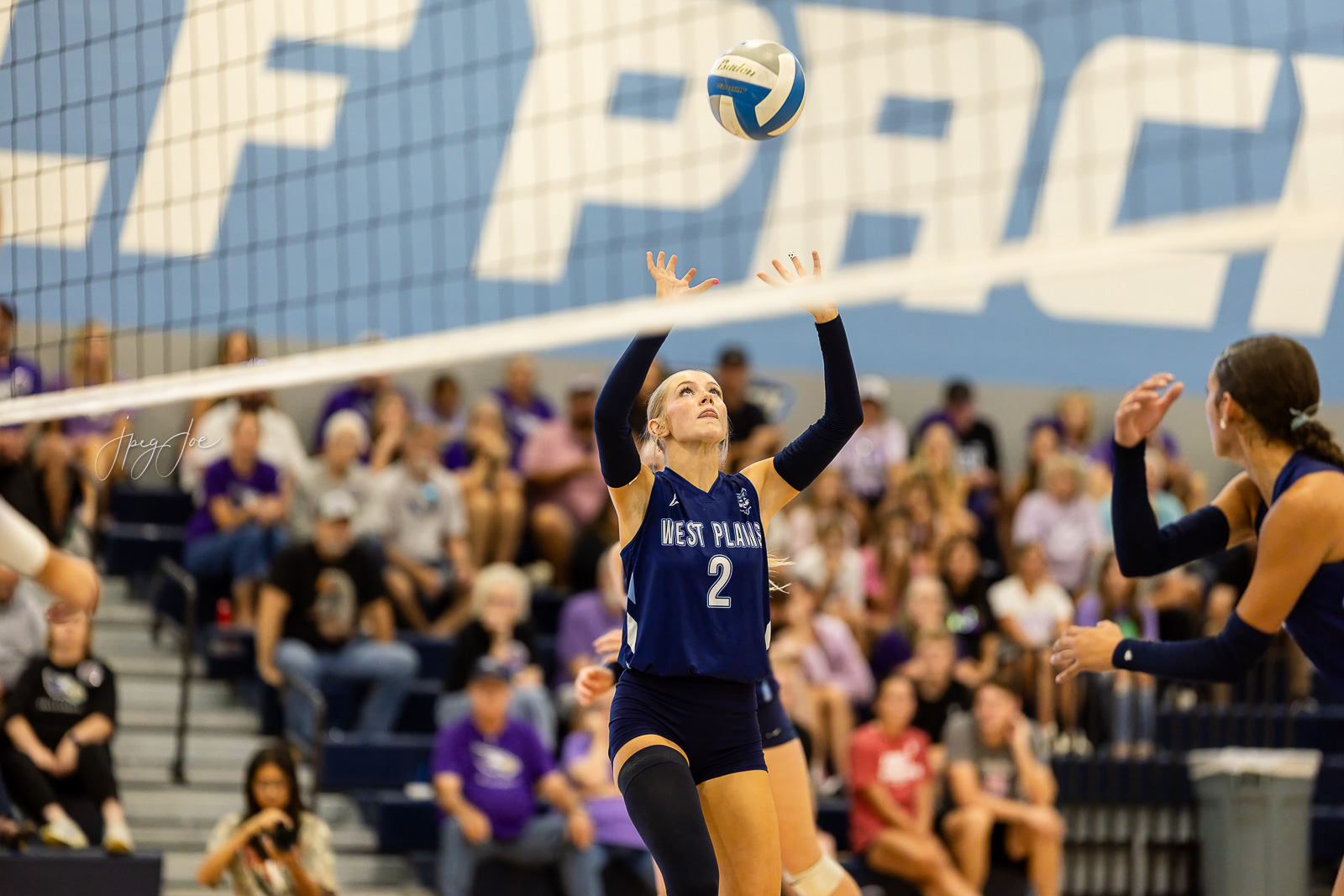 canyon-vs-west-plains-jpegJoe-volleyball-pps-35 | Press Pass Sports West Plains player #2 in blue jersey sets the ball at the net during a volleyball match, with a cheering crowd in the bleachers behind.