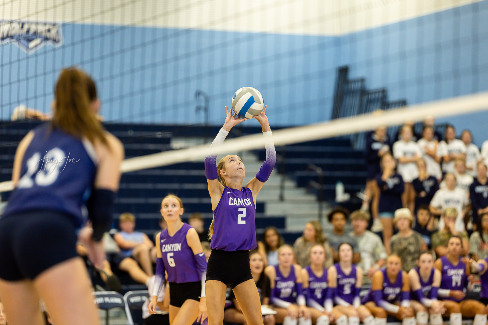 canyon-vs-west-plains-jpegJoe-volleyball-pps-38 | Press Pass Sports Setter in purple jersey number 2 sets the volleyball over the net during a match, with teammates and a cheering crowd in the gym.