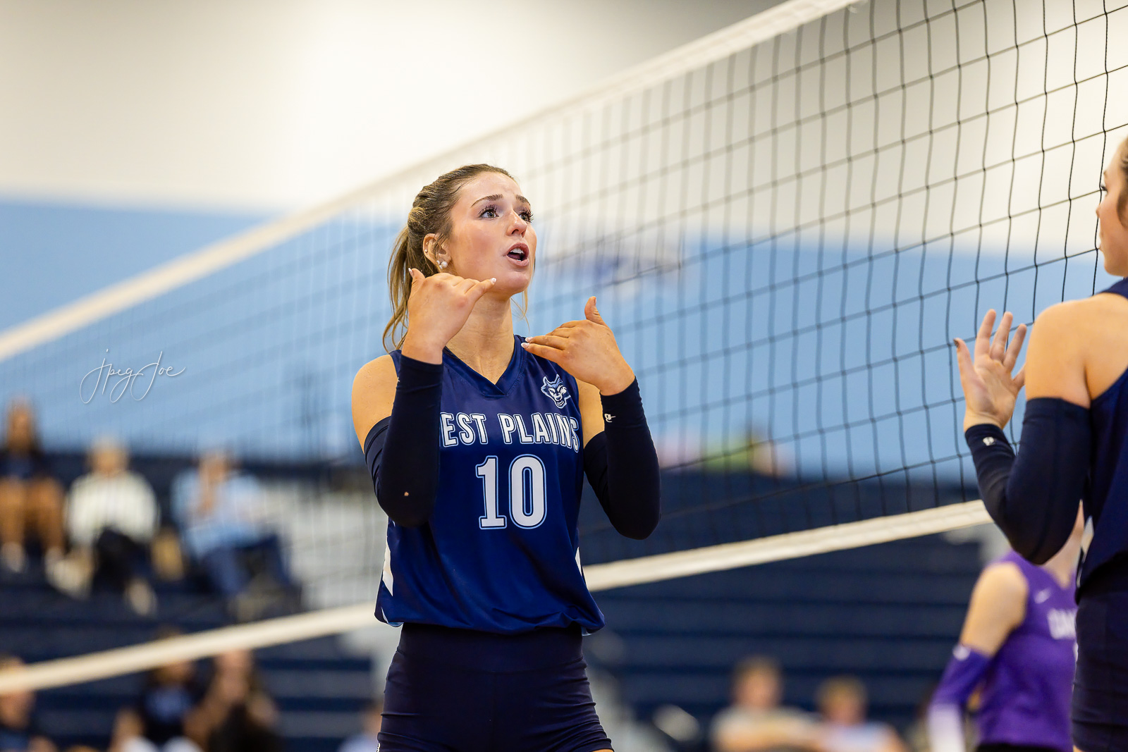 canyon-vs-west-plains-jpegJoe-volleyball-pps-39 | Press Pass Sports Volleyball player in a blue jersey number 10 at the net, signaling with her hands during a match in a gym.