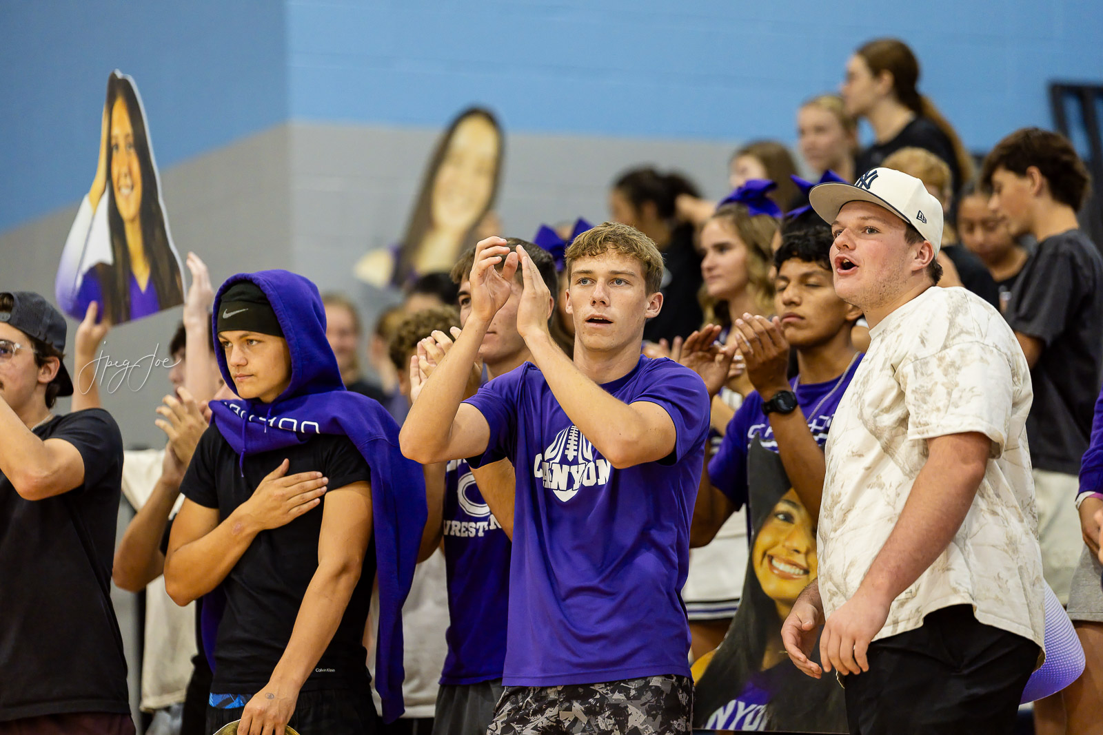 canyon-vs-west-plains-jpegJoe-volleyball-pps-42 | Press Pass Sports A group of high school students at a gym cheering, with a central boy in a purple shirt clapping and others around him wearing purple apparel and masks/prints in the background.