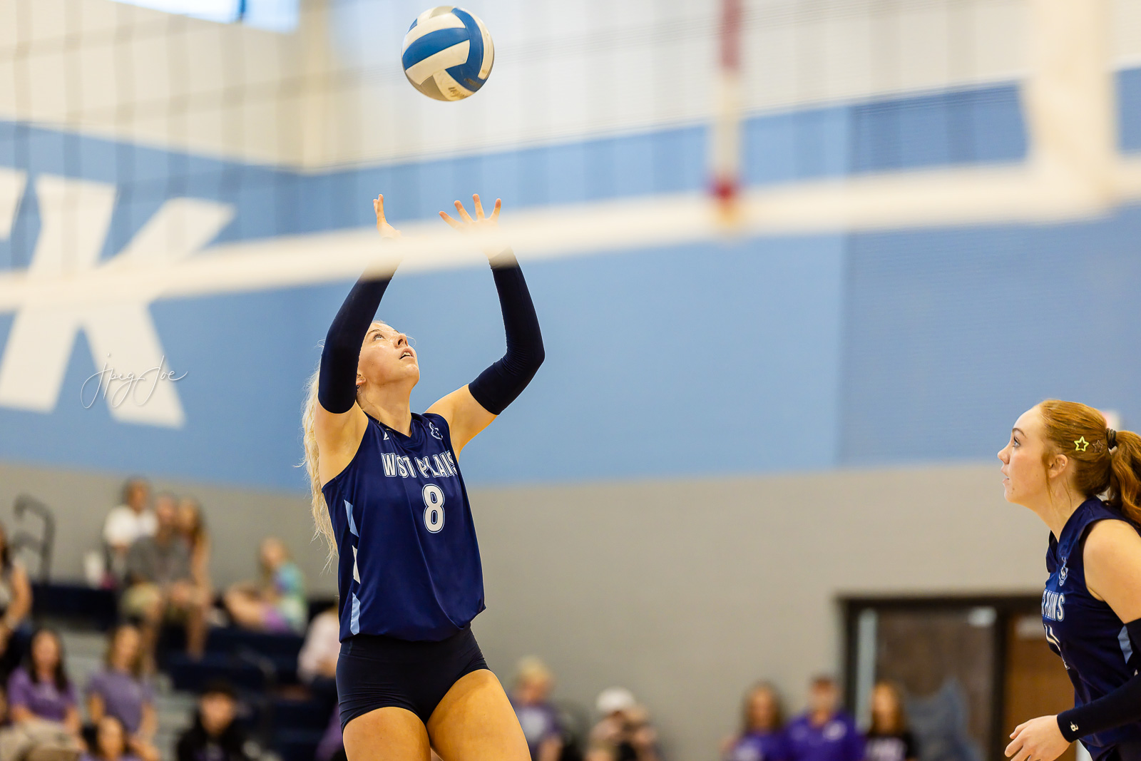 canyon-vs-west-plains-jpegJoe-volleyball-pps-44 | Press Pass Sports Volleyball player in a dark blue jersey (#8) jumps to set the ball over the net in a gym, with another teammate watching from the right and spectators in the background.