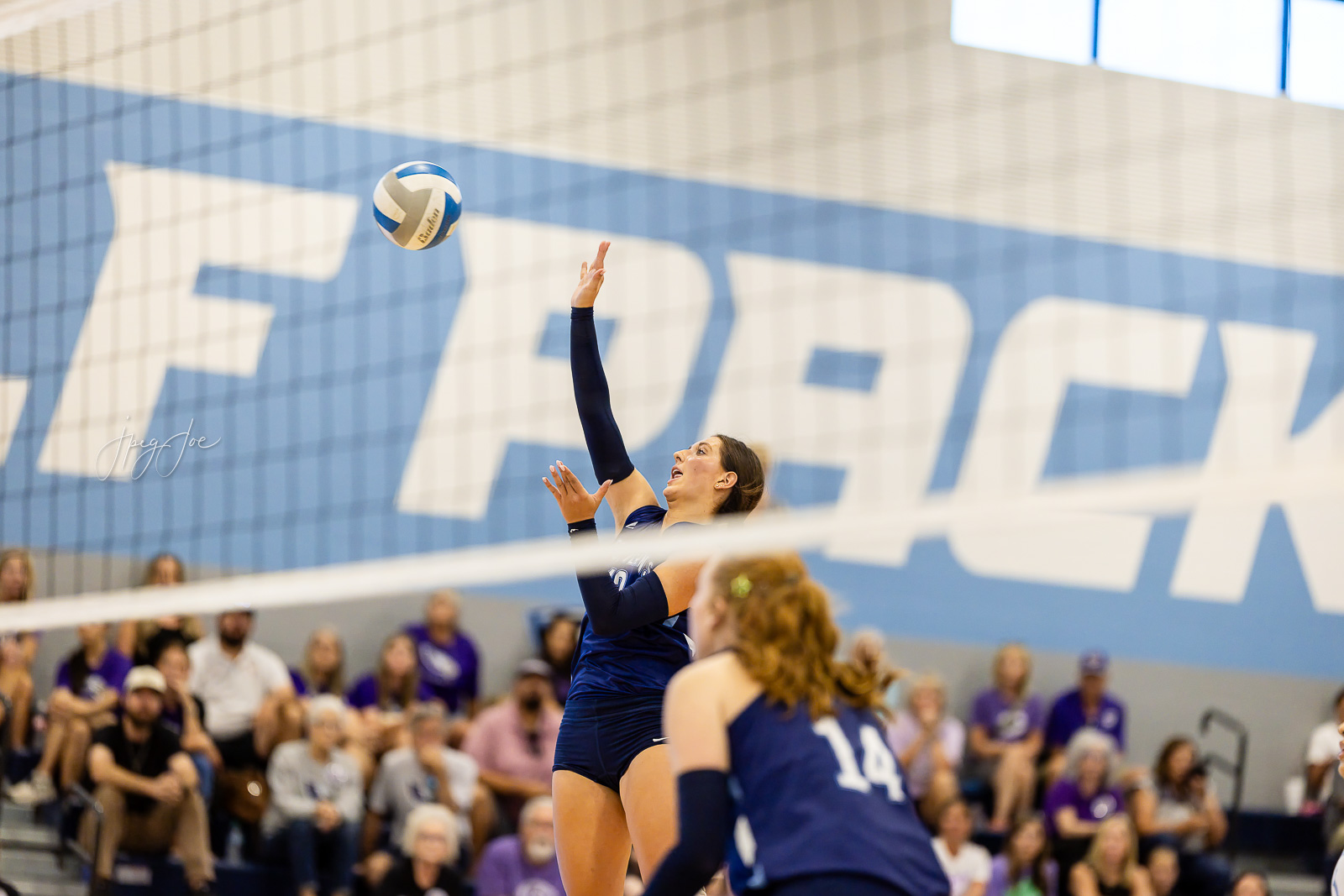 canyon-vs-west-plains-jpegJoe-volleyball-pps-45 | Press Pass Sports Female volleyball player in a navy uniform jumps to spike the ball over the net as spectators watch from the bleachers.
