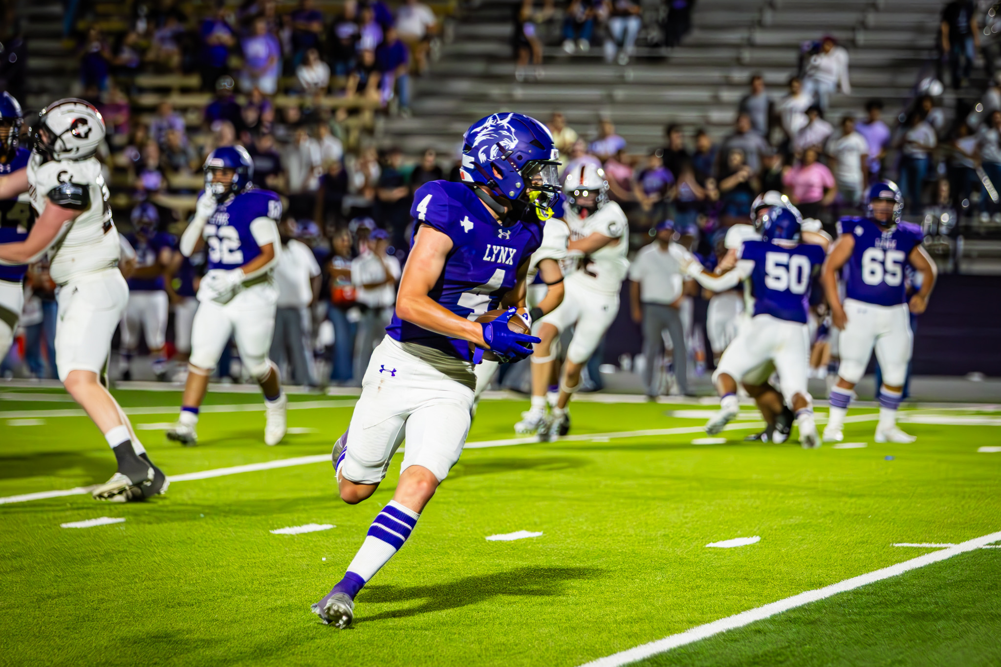 Football player in purple Lynx jersey (#4) runs with the ball on a bright green field; defenders and a crowded sideline in the background.