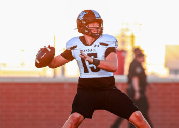 Football quarterback in white jersey (#13) holding ball, about to pass on a sunny sideline field.