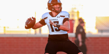 Football quarterback in white jersey (#13) holding ball, about to pass on a sunny sideline field.